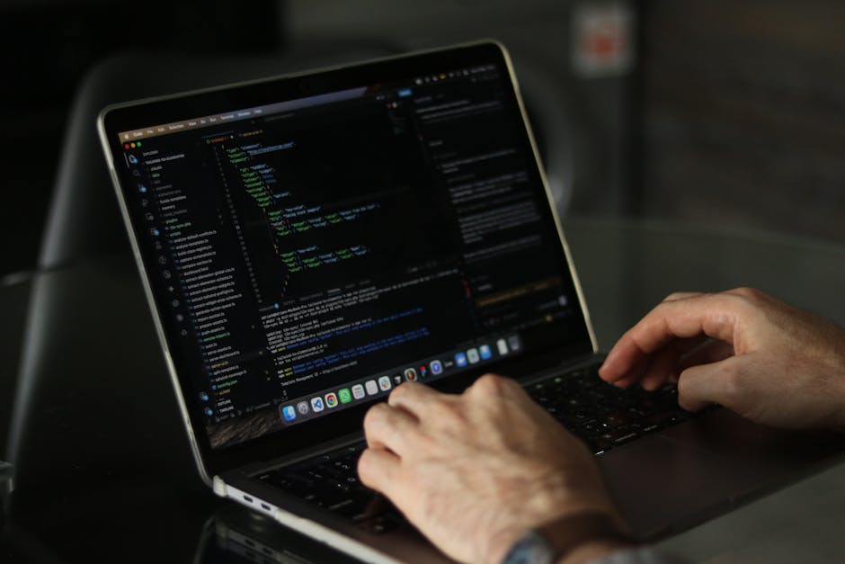 Close-up of a person's hands typing on a MacBook with a dark-themed coding editor open on the screen, displaying lines of green, blue, and white code. The laptop is placed on a black surface, and the 