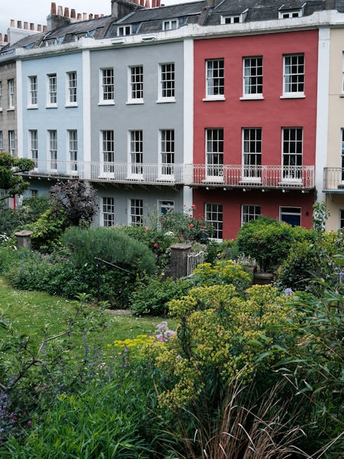 A multi-story period property with a facade painted in pastel blue and pink, featuring multiple white-framed sash windows. In the foreground, a lush garden with a variety of plants, including flowerin